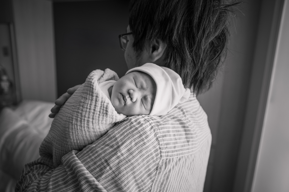 Black and white photo of father holding baby on his shoulder. The photo is taken from behind the father so you see the baby's face but not the father.