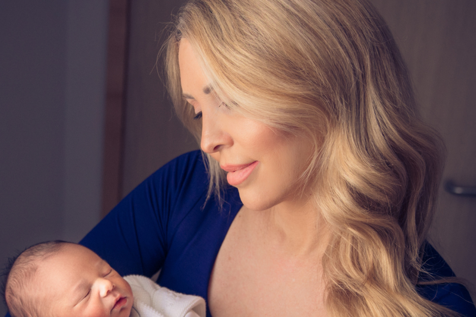 A woman with long, wavy blonde hair wearing a navy blue dress lovingly holds a sleeping newborn baby dressed in a white knitted outfit. The woman gazes down at the baby with a soft smile, standing in a warmly lit indoor setting with a wooden door in the background.