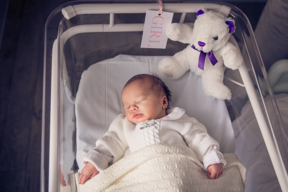 baby sleeps in bassinet. next to the baby is a white teddy with a purple bow around it's neck and a tag that says girl in capital pink letters.