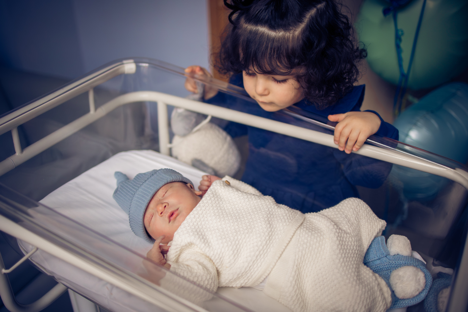 Sibling stares at newborn sibling sleeping in bassinet.
